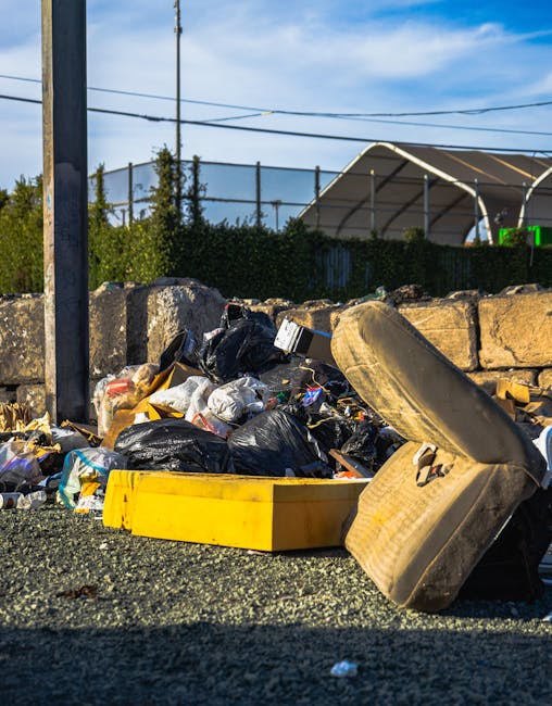 A piled heap of household rubbish and debris is located on a gravel surface next to a stone wall, with black garbage bags and loose waste materials visible, including a large, partially crushed beige sofa cushion leaning against the refuse. A bright yellow plastic container lies in front of the trash, which consists of mixed waste such as plastic packaging, paper, and other household refuse. In the background, there are green hedges, a metal fence, and an industrial or commercial building with a curved roof, suggesting the scene takes place outdoors near a property boundary or service area. Overhead, power lines and a utility pole are visible against a partly cloudy sky, and the overall environment appears to be an area designated for rubbish disposal or clearance, consistent with services offered by companies like House Clearance Croydon.