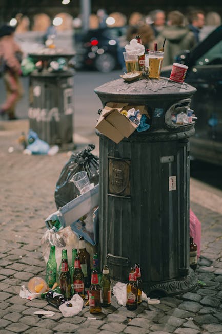 A piled heap of household rubbish and debris is located on a gravel surface next to a stone wall, with black garbage bags and loose waste materials visible, including a large, partially crushed beige sofa cushion leaning against the refuse. A bright yellow plastic container lies in front of the trash, which consists of mixed waste such as plastic packaging, paper, and other household refuse. In the background, there are green hedges, a metal fence, and an industrial or commercial building with a curved roof, suggesting the scene takes place outdoors near a property boundary or service area. Overhead, power lines and a utility pole are visible against a partly cloudy sky, and the overall environment appears to be an area designated for rubbish disposal or clearance, consistent with services offered by companies like House Clearance Croydon.