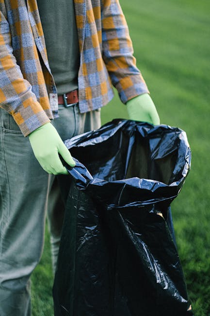 A person wearing a plaid shirt with shades of orange, blue, and grey, along with green gloves, is holding open a black plastic rubbish bag outdoors on a grassy area. The individual appears to be preparing or collecting waste, with a focus on the bag itself which has a shiny, slightly crinkled texture. The person's torso and hands are visible, but their face is not shown. In the background, there is a blurred expanse of green grass and possibly some shrubbery or trees, indicating a park or garden setting. The scene suggests an activity related to waste disposal or clearance, aligning with the context of rubbish removal services provided by companies like House Clearance Croydon, serving purposes such as independent collection or private disposal outside traditional municipal waste systems.
