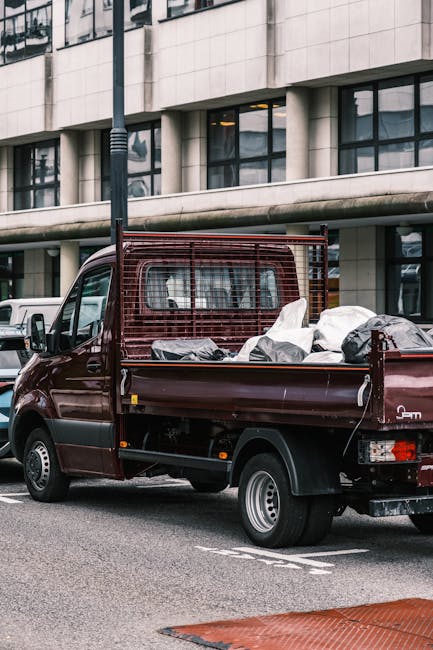 A burgundy flatbed truck parked on a city street in front of a modern office building with large glass windows. The truck's cargo area is filled with various bags and loose rubbish, including black plastic bags and white sacks, indicating it is being used for waste collection or clearance. The truck features a protective metal grid behind the cab and side mirrors positioned to the front. The surrounding environment includes a paved road with a designated parking area, a streetlamp near the vehicle, and a section of a red brick pavement in the foreground. The scene suggests an on-site waste removal operation, possibly managed by a private company such as House Clearance Croydon, aimed at alternative rubbish handling or bulk waste clearance outside traditional municipal collection services.