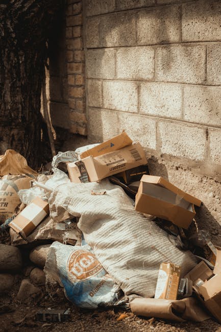 A pile of mixed waste materials is accumulated against a weathered brick wall, located outdoors on a narrow, uneven dirt surface, with some parts of the debris partially resting on small stones. The waste includes several flattened cardboard boxes of varying sizes, some with visible barcode labels, and beige and brown packaging with printed logos or text. There is a large, white textured sack partially covering the cardboard, possibly used for waste collection, and a blue plastic bag with orange logos or markings. The background features a light-colored brick wall with a rough, aged finish, and an adjacent darker brick section on the left side, with a small, partially visible tree trunk or post nearby. The scene appears to depict an informal rubbish collection or on-site waste disposal area, which House Clearance Croydon could assist with through private rubbish removal services. The lighting is natural, and the overall setting suggests a typical outdoor rubbish accumulation site requiring professional clearance. Throughout, the focus is on the waste materials and their immediate environment, relevant to waste handling and rubbish removal practices.