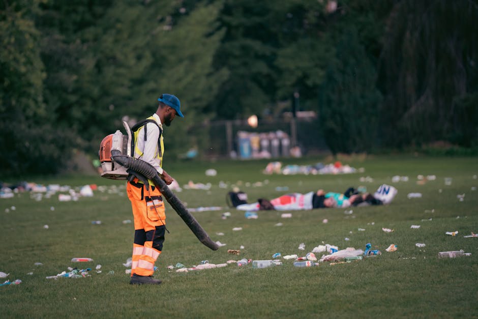 A worker in high-visibility orange trousers with reflective stripes, a grey jacket, and a blue cap is using a leaf blower to clear debris, including plastic bottles, paper, and other litter, from a grassy park area. The worker is carrying a backpack-style vacuum or blower and standing on a well-maintained green lawn. In the background, two people are lying on the grass among scattered rubbish, with trees and shrubs surrounding the area, and a few trash bins are visible further back. The scene appears to be an outdoor cleanup operation, possibly related to environmental or waste management services, with natural light illuminating the scene and highlighting the accumulation of litter that House Clearance Croydon might handle through alternative waste disposal methods.