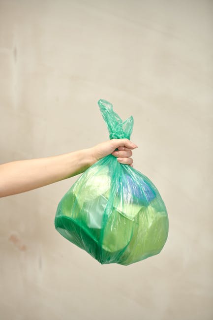 A person's arm and hand extending from the left side of the image are holding a green plastic rubbish bag that appears to contain waste, with the top of the bag gathered and twisted into a knot. The bag is semi-transparent, revealing some lighter and darker areas of waste material inside, and has a slight sheen reflecting light. The background is a plain, neutral beige wall, and the scene is evenly lit without shadows. This image relates to rubbish collection or waste disposal, reflecting an aspect of independent waste handling often associated with private rubbish removal services like those offered by House Clearance Croydon, especially in contexts such as garden or household clearance.
