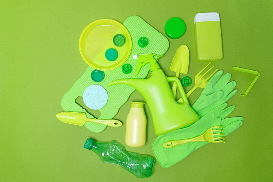 A collection of bright green home cleaning tools and accessories arranged on a flat surface, including a rubber watering can, a pair of rubber gloves, a plastic spray bottle, a small bottle of cleaning solution, a scrub brush, a sponge, a plastic scraper, a small plastic scoop, and a few small caps or lids. The background is a vibrant green, matching the color of most items, creating a cohesive visual effect. The arrangement appears to be organized but casual, with objects overlapping slightly, suggesting preparation for cleaning or waste collection. The image illustrates domestic cleaning equipment typically associated with household maintenance or rubbish removal services provided by companies like House Clearance Croydon, and highlights the focus on eco-friendly or color-themed cleaning supplies suitable for on-site clearance tasks involving garden or home waste. The overall scene emphasizes a neat, practical setup of cleaning tools ready for use or disposal.