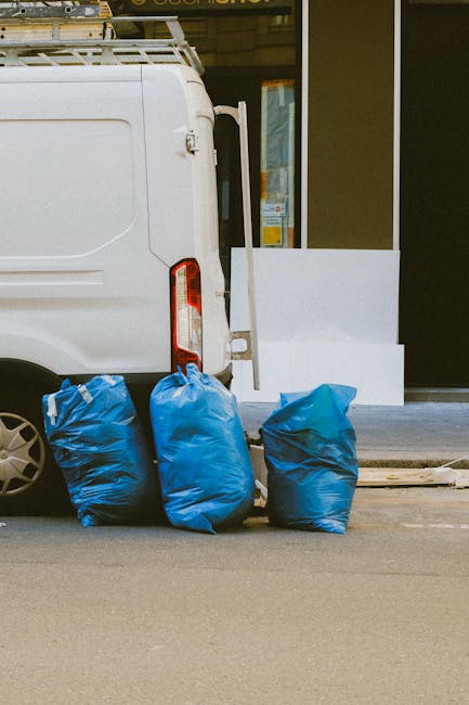 In the foreground, three large blue rubbish bags made of thick, woven plastic material are positioned on a paved street surface adjacent to the rear of a white commercial van. The bags are filled with waste and appear slightly bulging, with one bag leaning to the side. The van's rear door and taillight are visible on the left side of the image, showing a clean, modern design with a red and white taillight assembly. Behind the bags, part of a building's exterior is visible, including a large glass window or door framed in black, with a concrete step or platform leading up to it. The lighting suggests natural daylight, and the scene appears to be part of a private or commercial rubbish collection effort, typical of on-site waste handling services like those offered by House Clearance Croydon. The overall environment is urban and tidy, with minimal clutter, reflecting a professional rubbish removal process aimed at quick and efficient waste clearance, possibly related to independent collection services rather than local authority disposal.